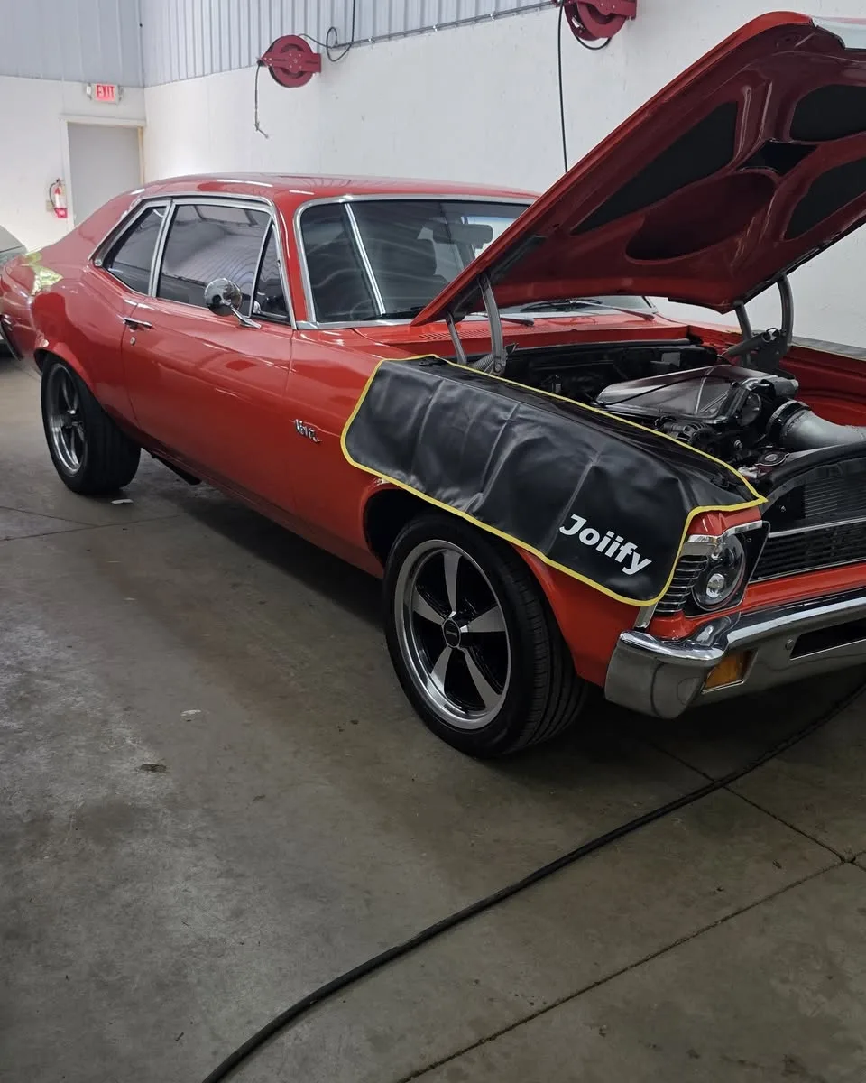 A red Chevy Nova with its hood up and a fender cover on, in the shop’s bay